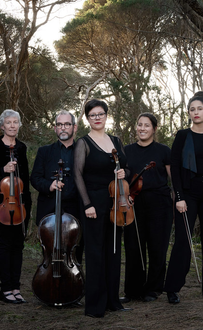 A group of musicians hold their instruments surrounded by bushland. The musicians wear wear head to toe black. 