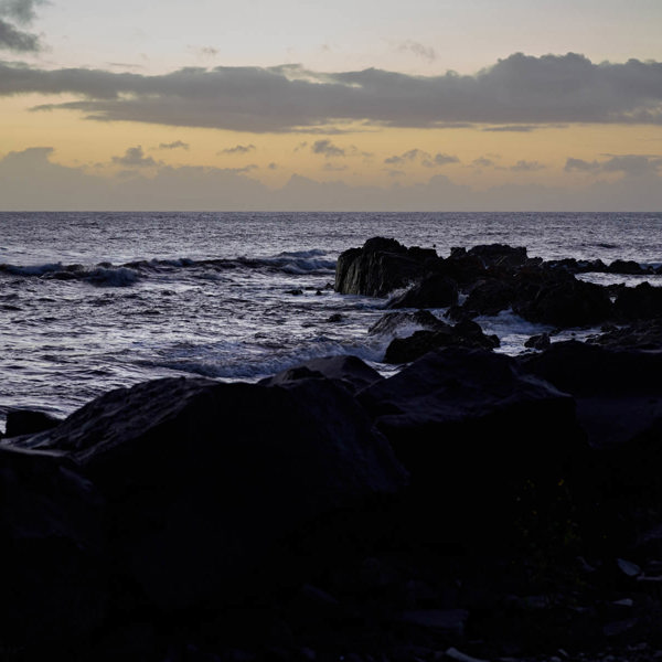 A yellow and blue sunset, the wild ocean is in the foreground but is covered by the shadow 