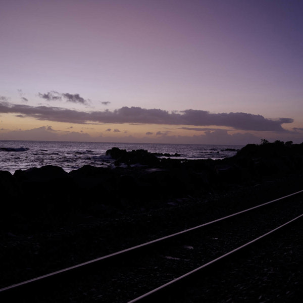 A light purple and yellow sunset, outlines of rocks and what looks to be a railroad are in the foreground but are covered by the shadow 