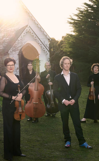 A group of people hold musical instruments standing on the grass in front of an old white small church, this church is surrounded by bushland