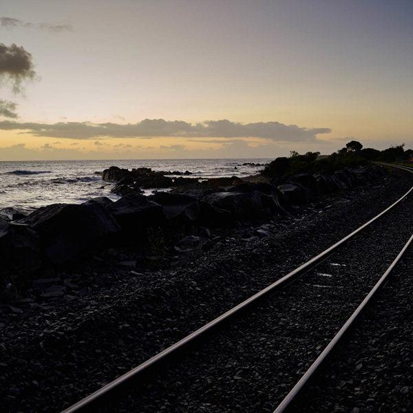 A light yellow sunset, outlines of rocks and what looks to be a railroad are in the foreground but are covered by the shadow 
