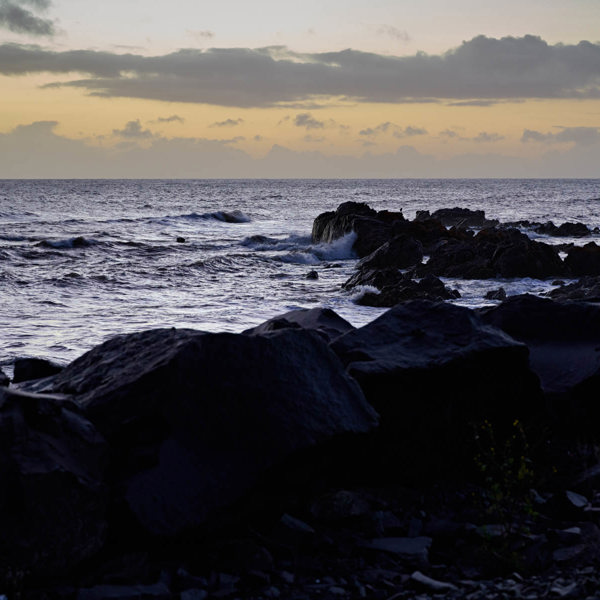 A yellow and blue sunset, the wild ocean with rocks on the shore is in the foreground but is covered by the shadow