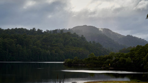 there is a glistening body of water, under beautiful sunlight, a wild mountainous backdrop and a yellow grass spot of land in the forground