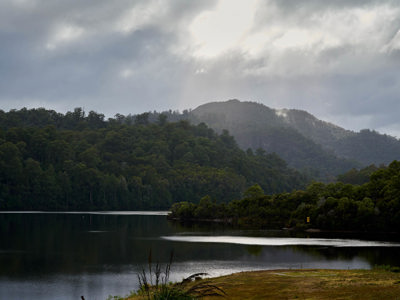 there is a glistening body of water, under beautiful sunlight, a wild mountainous backdrop and a yellow grass spot of land in the forground