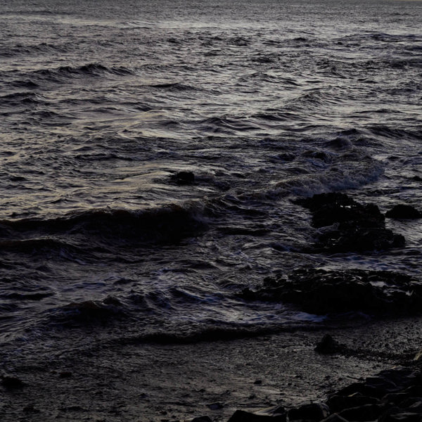 A dark ocean with only the waves and rock shadows recognisable.