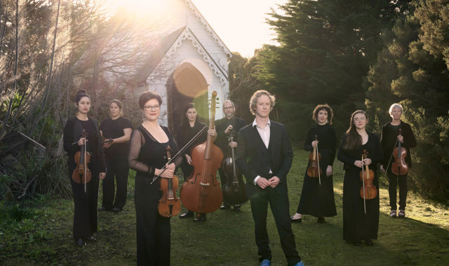 A group of people hold musical instruments standing on the grass in front of an old white small church, this church is surrounded by bushland